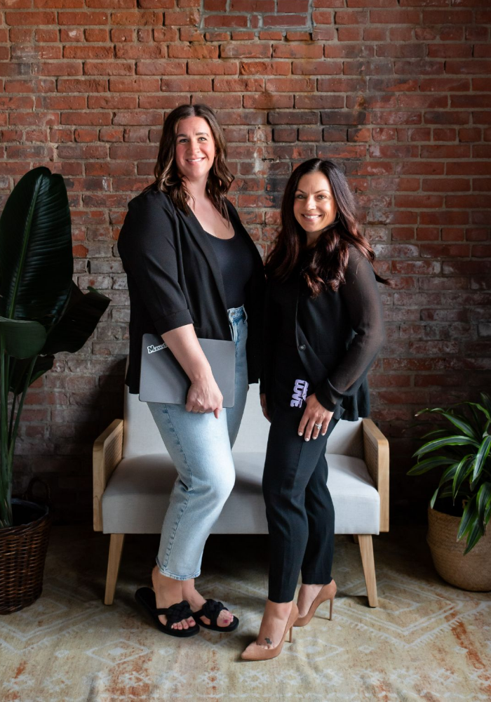 Two women founders of FiZZY, a brand offering patented hair claws for women, standing and posing together in front of a brick wall.